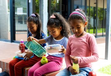 Three students share lunch, showing joy and diversity. An African American girl in pink top, and two Hispanic girls, one in blue, other in pink, chat happily, unaltered