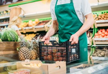 partial view of shop assistant holding basket with fruits in grocery shop