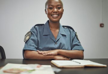 Happy, smile and portrait of a female police officer sitting by a desk in the office writing report.