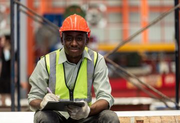 Happy male african american workers wear red helmet working take notes on paperwork sitting in factory. Heavy Industrial manufacture workplace.