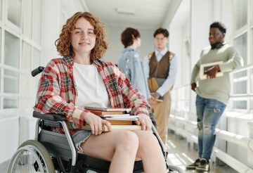 Smiling teenage girl with physical disability in casualwear sitting in wheelchair in college corridor against group of classmates